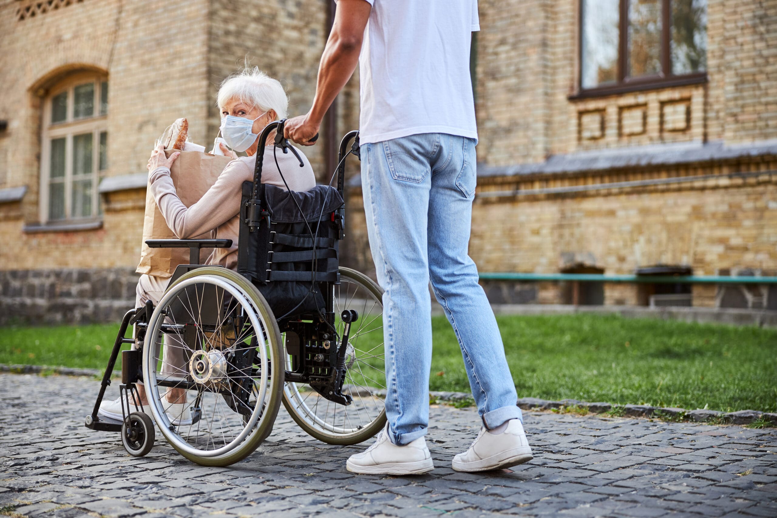 Elderly lady with disability holding paper bag with purchases and turning back while young person pushing her wheelchair